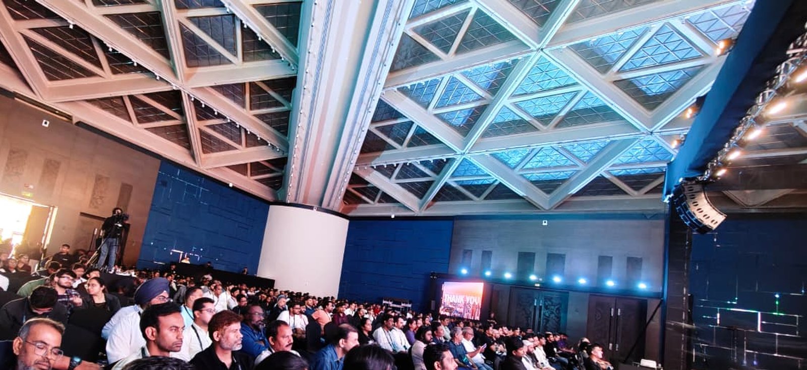 Wide shot of the packed Developer Days Bengaluru auditorium, audience seated, venue's glass-triangle ceiling visible.